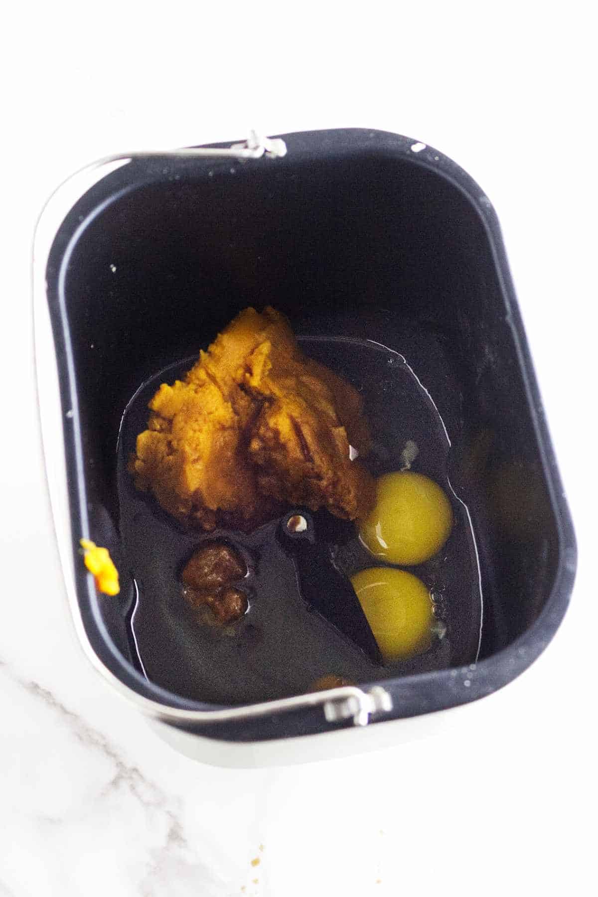 A bread machine pan containing pumpkin puree, two cracked eggs, and other wet ingredients sits on a white surface, ready to be mixed into delicious bread machine pumpkin bread.