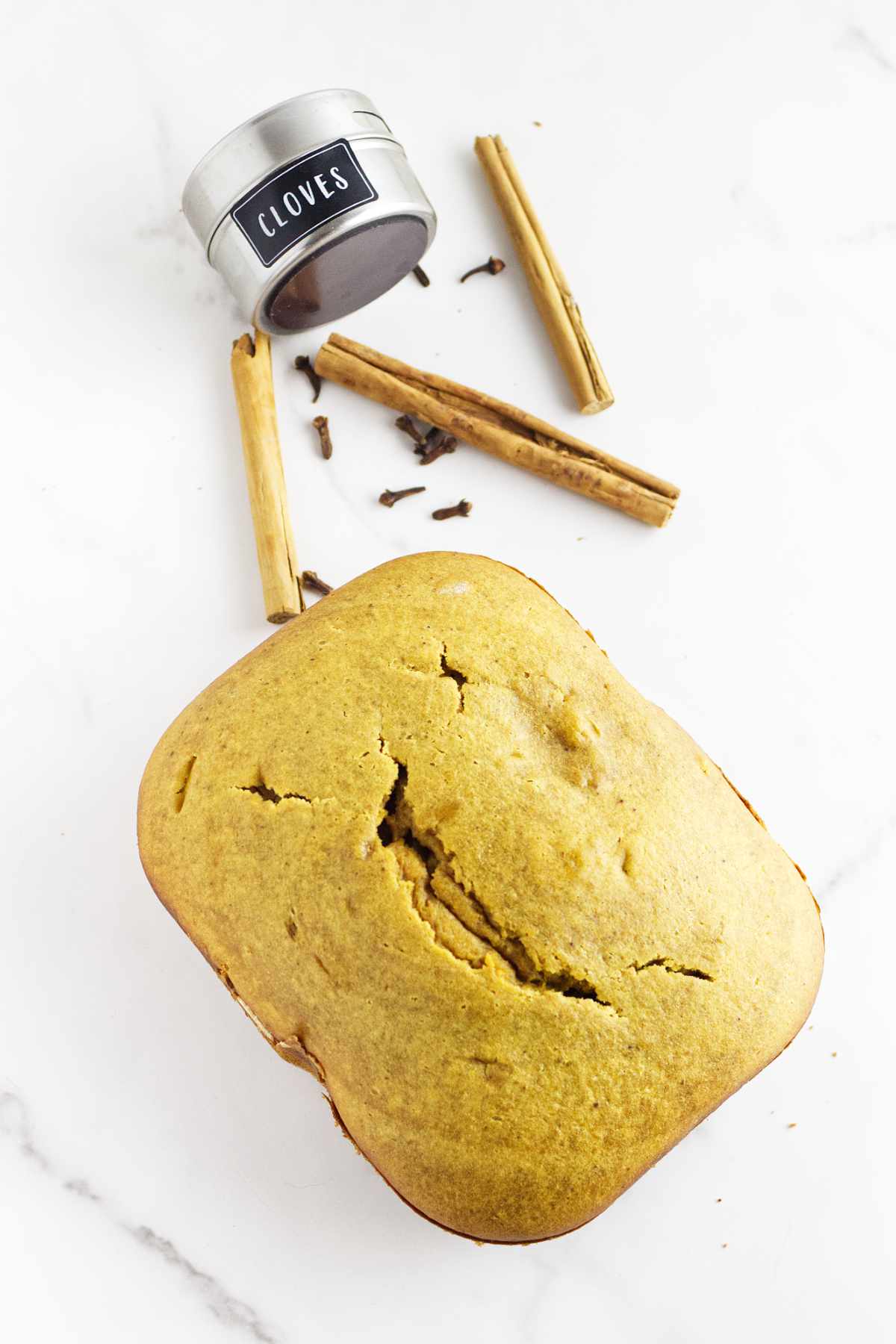 A loaf of bread machine pumpkin bread sits on a white surface next to whole cinnamon sticks, scattered cloves, and a small metal container labeled "Cloves" with its lid open.
