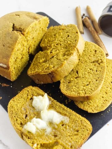 A loaf of bread machine pumpkin bread, partially sliced, sits on a black cutting board. Several slices are shown, with one spread with melted butter. Nearby are cinnamon sticks and a small jar labeled "cloves.