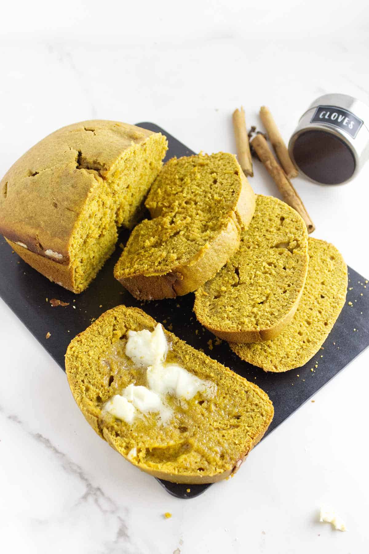 Sliced bread machine pumpkin bread on a black cutting board, with one slice spread with butter. Cinnamon sticks and a small container of cloves are in the background, all set on a white surface.