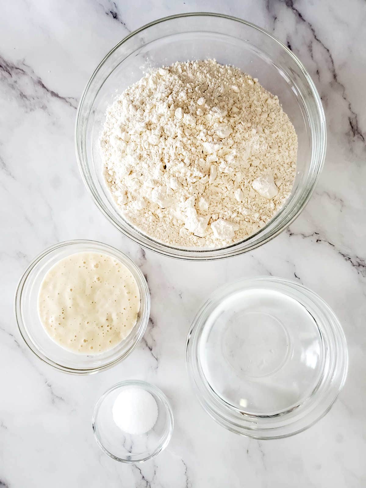 Four glass bowls on a marble surface showcase essential bread ingredients: flour, sourdough starter, water, and salt, all arranged for bread making.