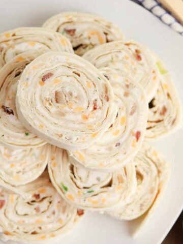 A plate of bacon ranch pinwheels filled with a creamy mixture of shredded cheese, bits of bacon, and green onions, sliced and arranged in a circular stack.