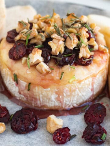 A round wheel of baked brie with cranberries, topped with chopped nuts and herbs, is surrounded by slices of bread and pear on parchment paper.