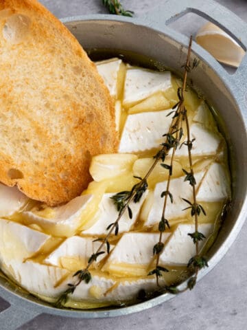 A slice of toasted bread is dipped into a round of baked brie with honey, topped with olive oil, garlic slices, and fresh thyme in a small metal dish.