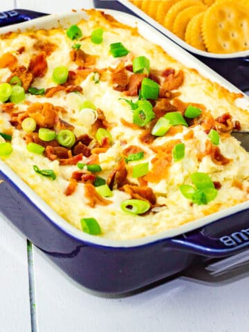 A blue casserole dish filled with creamy bacon ranch cheddar dip, topped with chopped green onions and crispy bacon bits, with a spoon inside. A tray of round crackers sits in the background.