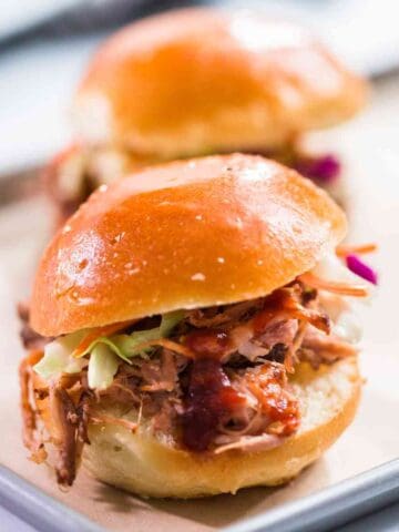 A close-up of BBQ pulled pork sliders with barbecue sauce, coleslaw, and a shiny brioche bun, placed on a tray with another slider blurred in the background.