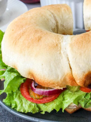 A large, ring-shaped bundt-wich party sub sits on a gray plate, filled with lettuce, tomato slices, pickles, and red onion. A bowl and stacked plates are blurred in the background.