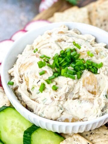 A white bowl filled with creamy caramelized onion dip topped with chopped green onions, surrounded by cucumber slices, radish slices, and crackers.