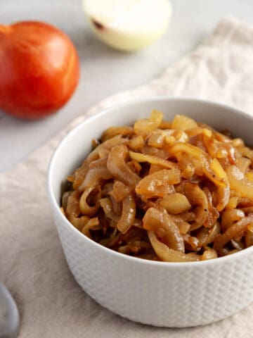 A white bowl filled with caramelized onions sits on a beige cloth, hinting at how to caramelize onions perfectly, with a whole and a half onion in the background and a spoon in the foreground.