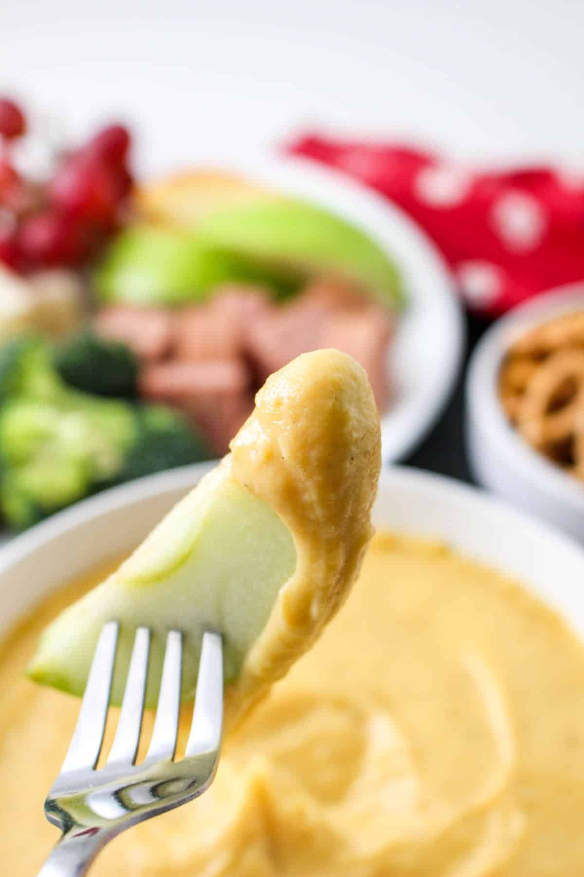 A fork holds a slice of green apple dipped in creamy cheddar cheese fondue, with bowls of broccoli, pretzels, fruit, and a red napkin blurred in the background.