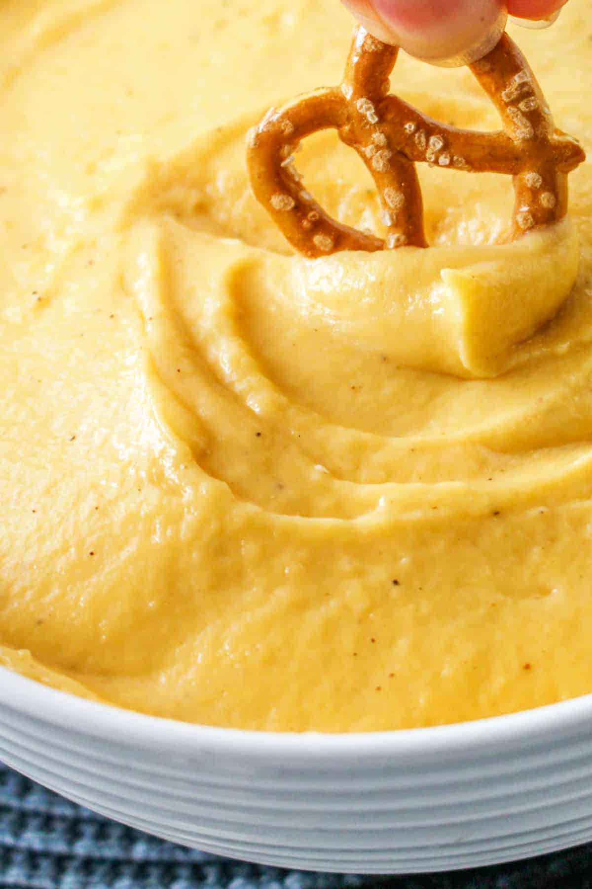 A hand dips a small salted pretzel into a creamy, yellow cheddar cheese fondue in a white bowl.