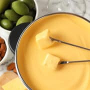 A pot of melted cheddar cheese fondue with two fondue forks holding cheese cubes, surrounded by green olives, almonds, grapes, and a glass of white wine on a marble surface.