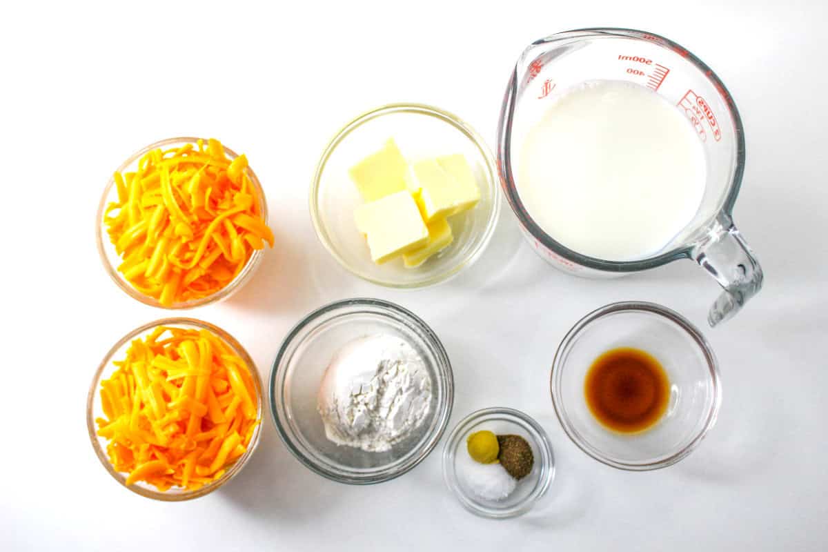 Overhead view of clear bowls containing cheddar cheese fondue ingredients—shredded cheddar cheese, butter, flour, milk in a measuring cup, salt, mustard, nutmeg, and vanilla extract—arranged on a white surface.