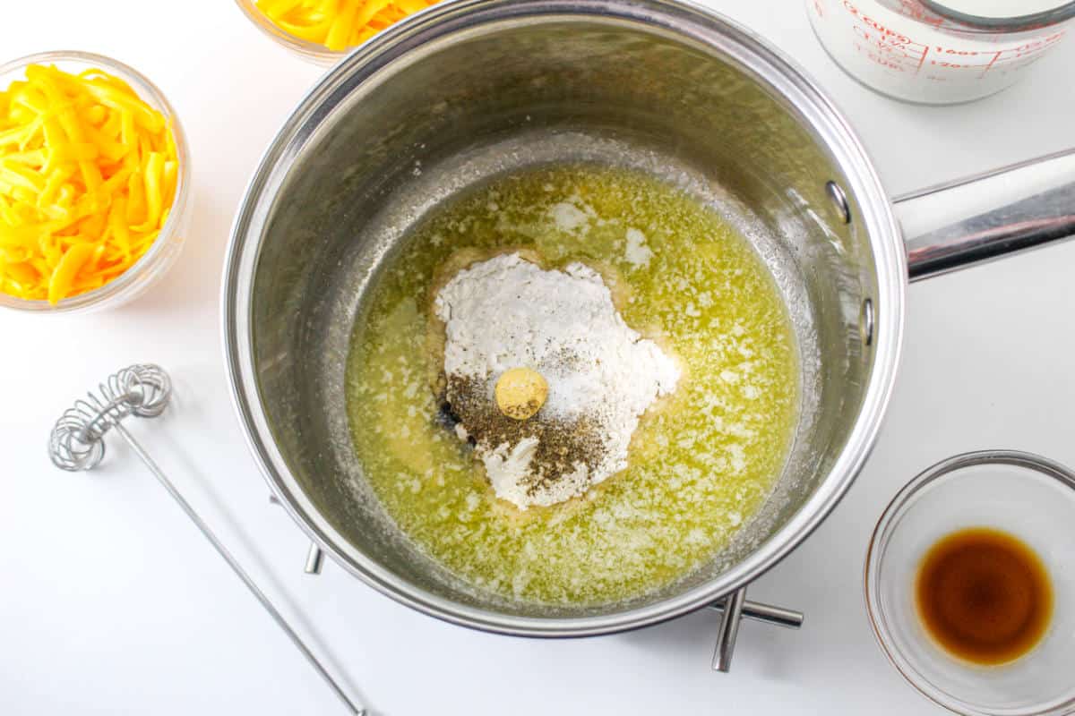 A saucepan with melted butter, flour, salt, and pepper, ready to be mixed for cheddar cheese fondue. Surrounding the pan are bowls of shredded cheese, milk, and vanilla, along with a whisk on a white countertop.