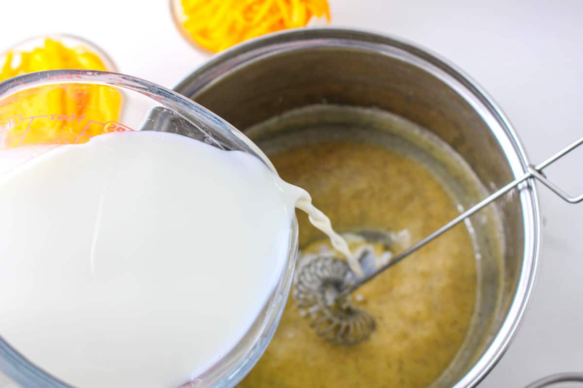 A glass measuring cup pours milk into a saucepan with a light brown mixture being whisked; small bowls of shredded cheddar cheese for fondue are visible nearby.