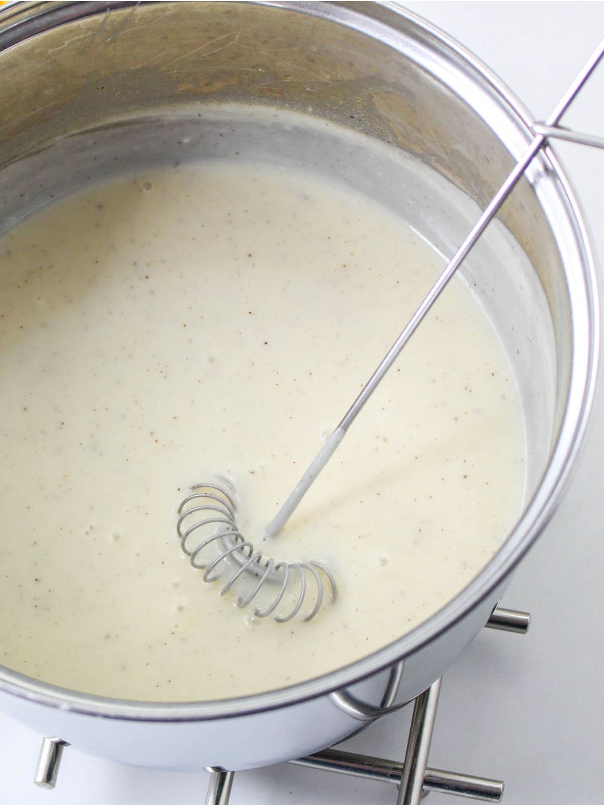 A metal whisk mixing creamy white cheddar cheese fondue with specks of seasoning in a stainless steel pot on a silver stove.