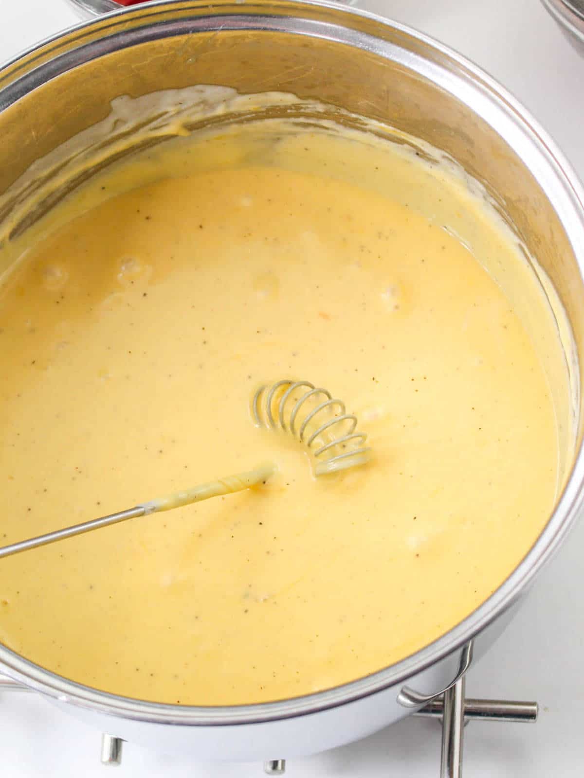A stainless steel pot filled with creamy yellow cheddar cheese fondue is being stirred by a metal whisk, with visible black pepper specks in the rich sauce.