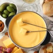 A pot of melted cheddar cheese fondue with two skewers holding bread cubes, surrounded by green olives, almonds, grapes, cheese slices, and pieces of bread on a marble surface.