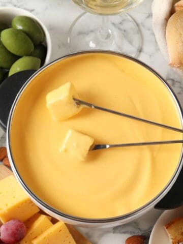 A pot of melted cheddar cheese fondue with two skewers holding bread cubes, surrounded by green olives, almonds, grapes, cheese slices, and pieces of bread on a marble surface.