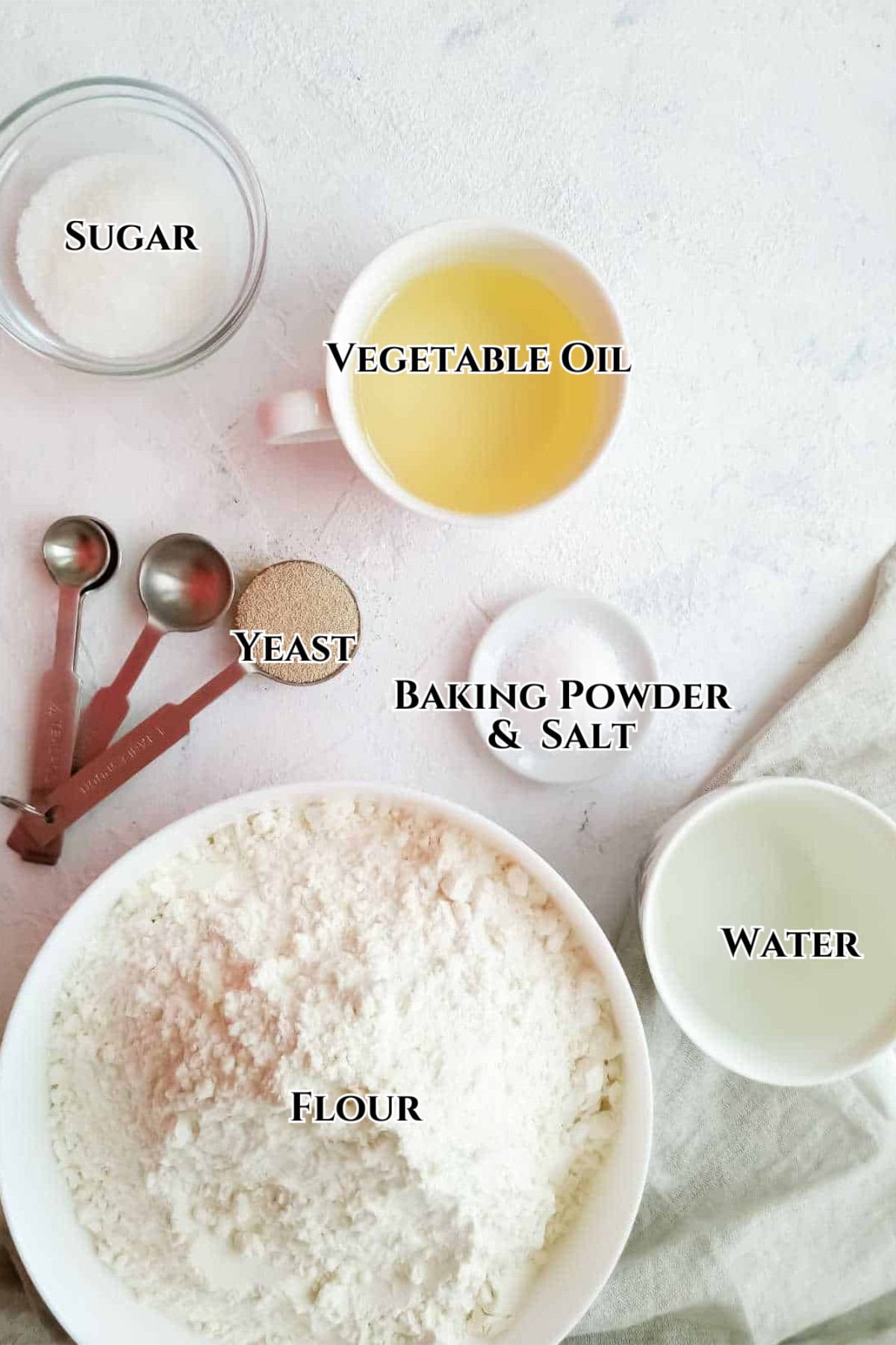 A top view of baking ingredients for Chinese steamed buns in bowls, labeled as sugar, vegetable oil, yeast, baking powder & salt, water, and flour, with measuring spoons and a light cloth on a white surface.
