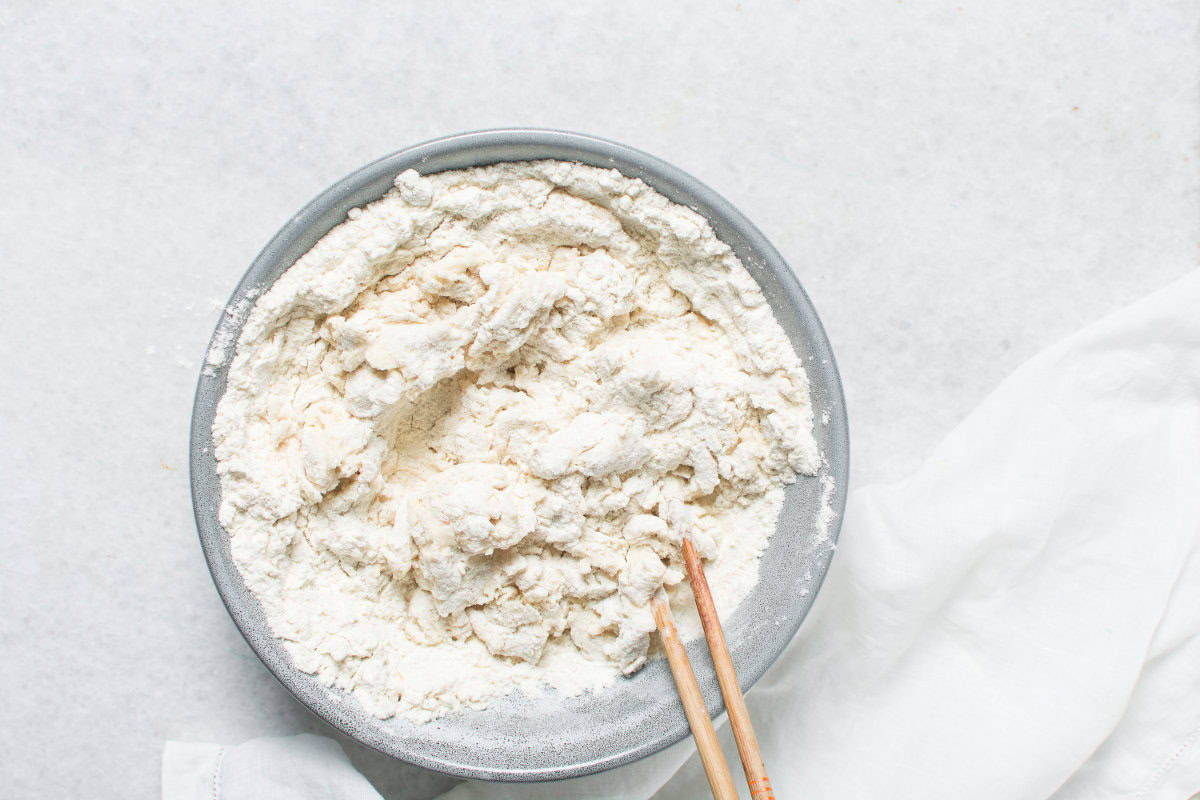 A gray bowl filled with flour and partially mixed dough for Chinese steamed buns, with a pair of wooden chopsticks resting in the bowl, placed on a white surface next to a white cloth.