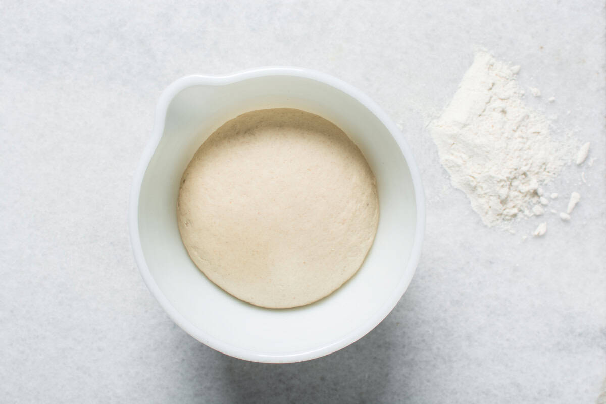 A ball of risen dough sits in a white bowl on a light surface, ready to be shaped into Chinese steamed buns, with a small pile of flour nearby.