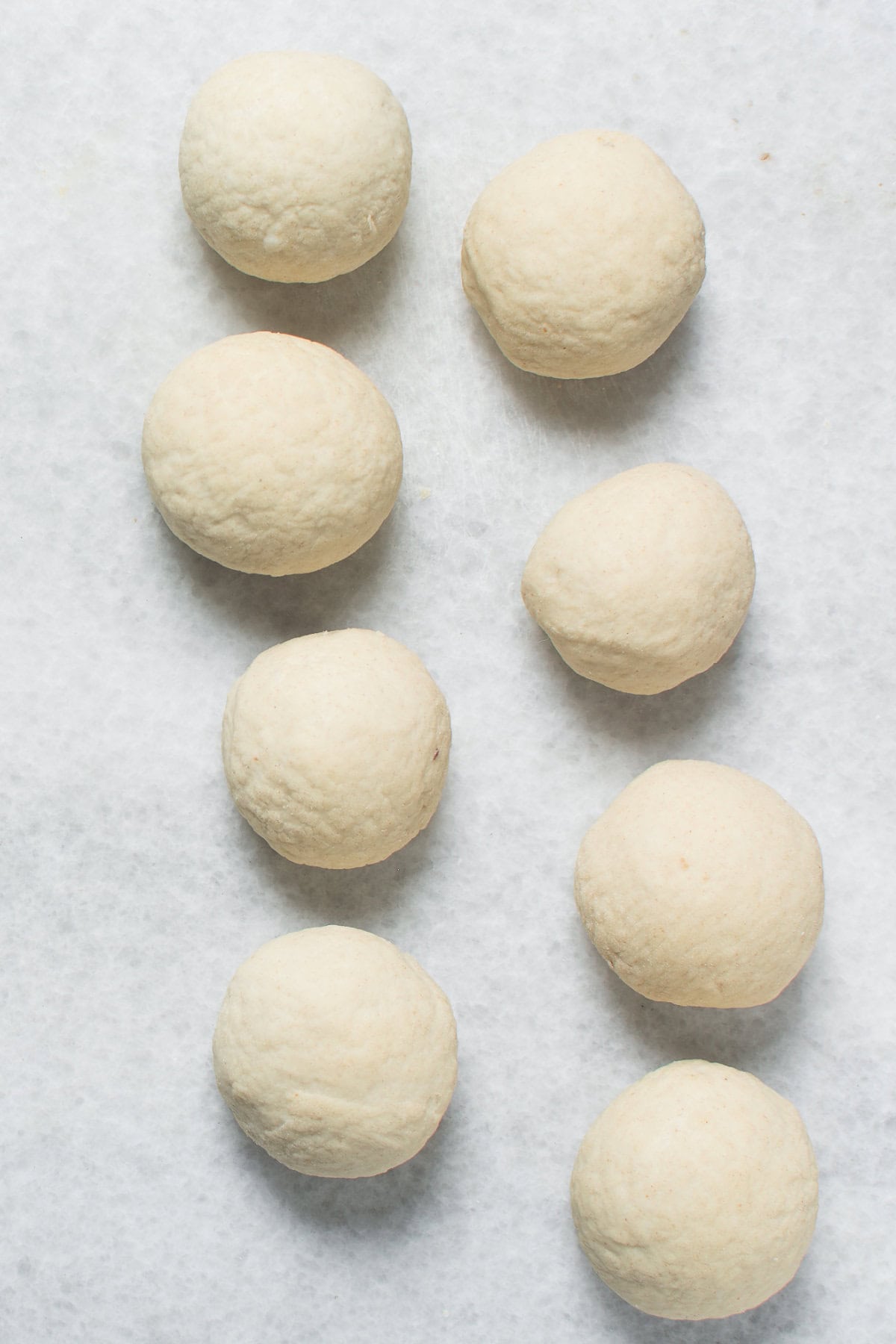 Eight round balls of dough, resembling chinese steamed buns, are arranged on a light-colored, textured surface in two uneven columns. The uncooked dough balls are evenly spaced apart.