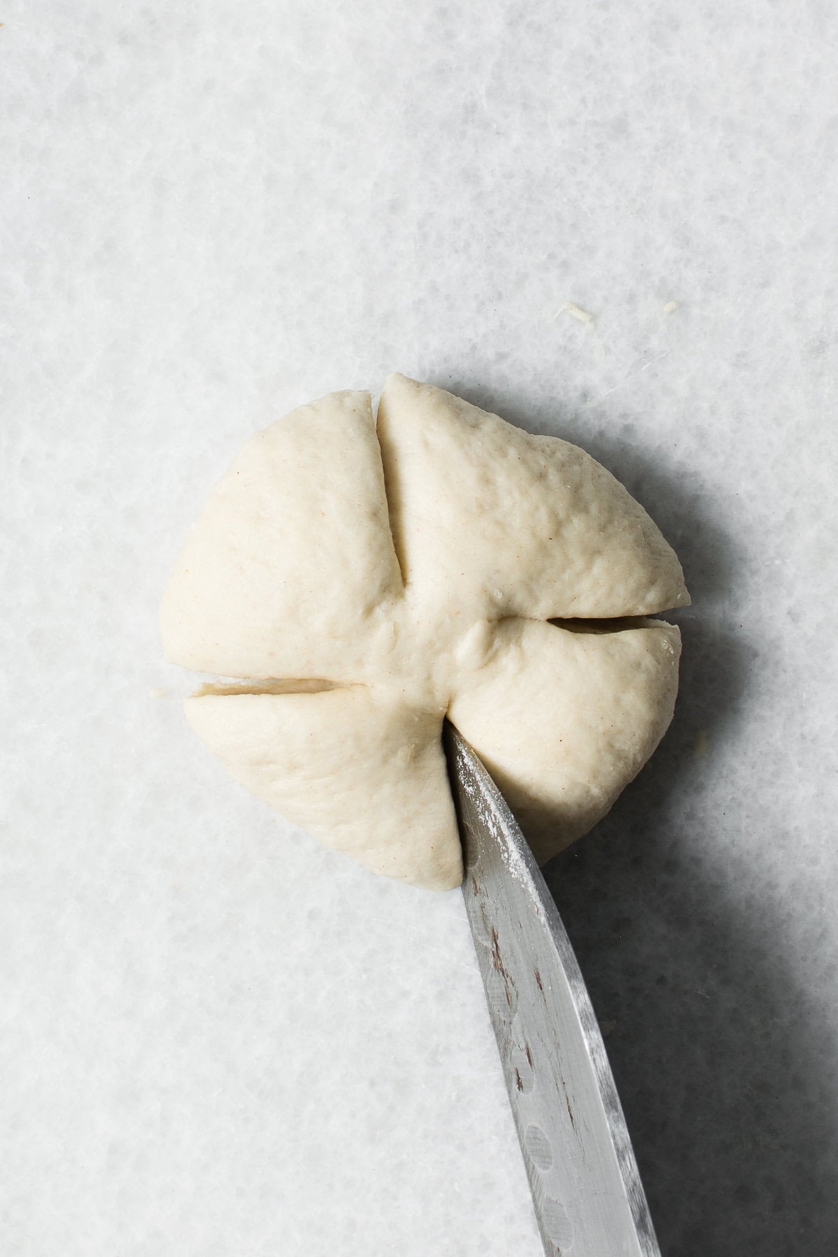 A ball of dough on a light surface being scored with a knife to create four sections on top, resembling the preparation for traditional Chinese steamed buns.