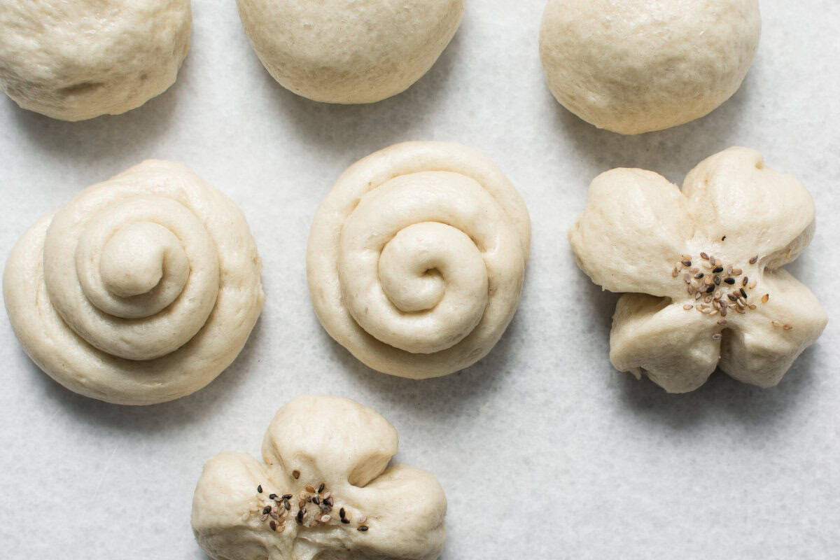 Six unbaked dough shapes on a white surface: two round balls, two spirals, and two flower shapes with small seeds or seasoning in the center, resembling traditional Chinese steamed buns.