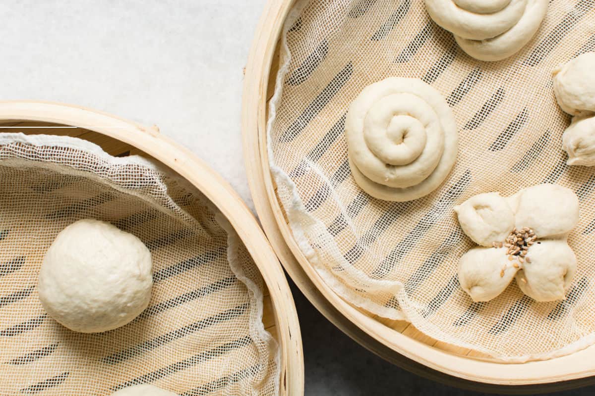 Two bamboo steamers contain chinese steamed buns on mesh liners: one holds a plain dough ball, while the other features a swirl and a flower-shaped bun with sesame seeds in the center.