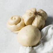 Three Chinese steamed buns in different shapes-a round bun, a flower-shaped bun topped with seeds, and a swirl-shaped bun-are arranged on a light surface beside a white cloth.