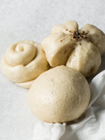 Three Chinese steamed buns in different shapes—a round bun, a flower-shaped bun topped with seeds, and a swirl-shaped bun—are arranged on a light surface beside a white cloth.