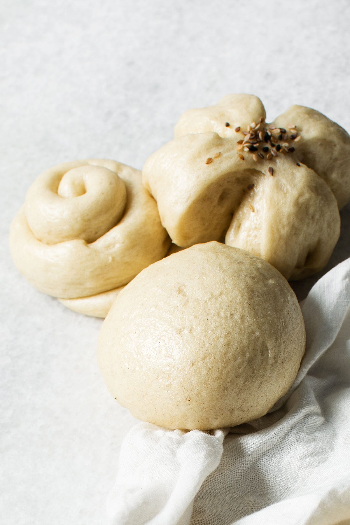 Three chinese steamed buns of different shapes—a round bun, a swirl, and a flower-shaped roll topped with seeds—are arranged on a light surface with a white cloth beside them.