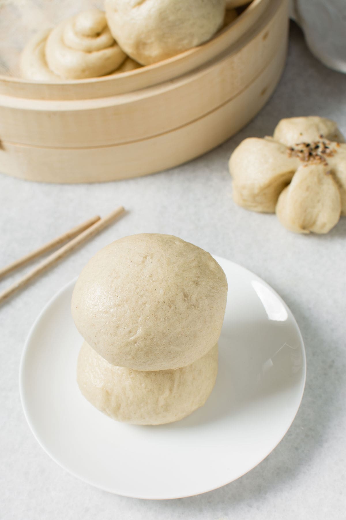 Two Chinese steamed buns are stacked on a white plate, with chopsticks beside them. In the background, more buns are visible in a bamboo steamer, and one flower-shaped bun rests on the table.
