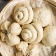 A bamboo steamer basket lined with cloth holds various shapes of uncooked Chinese steamed buns, showcasing round, spiral, and flower-shaped buns ready for steaming.