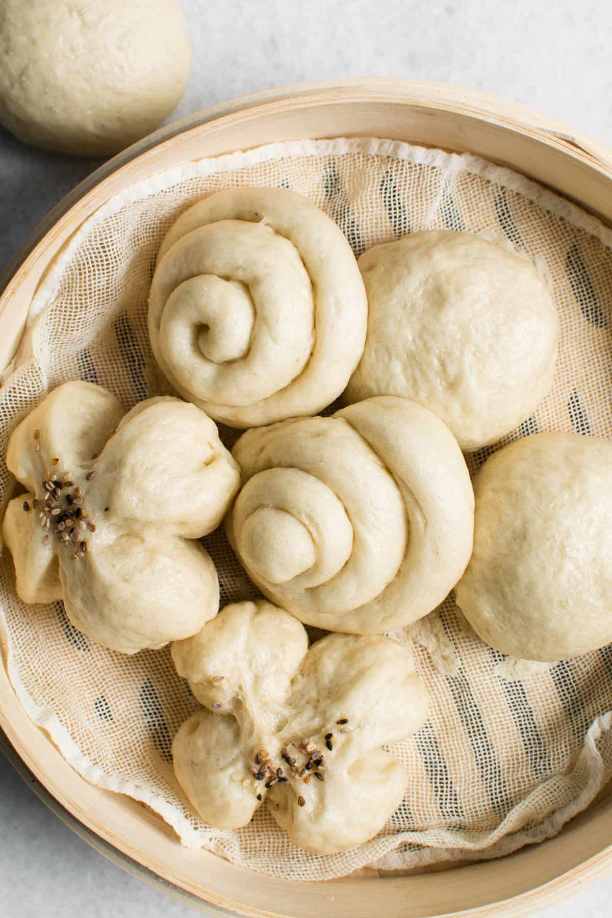 A bamboo steamer basket lined with cloth holds various shapes of uncooked Chinese steamed buns, showcasing round, spiral, and flower-shaped buns ready for steaming.
