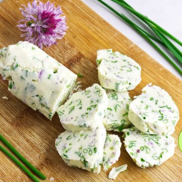 A log of fresh chive compound butter with chopped herbs is sliced on a wooden cutting board. Fresh chives and a purple chive flower are placed nearby for garnish.