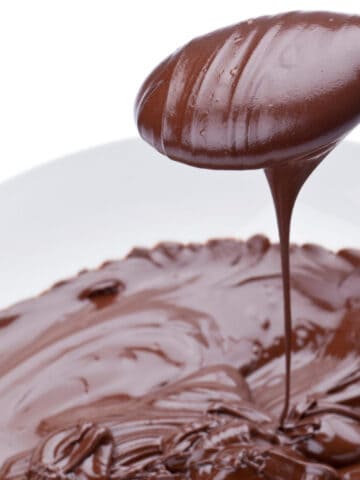 A spoon drizzles smooth, melted chocolate ganache over a plate with a thick layer of glossy chocolate filling against a white background.