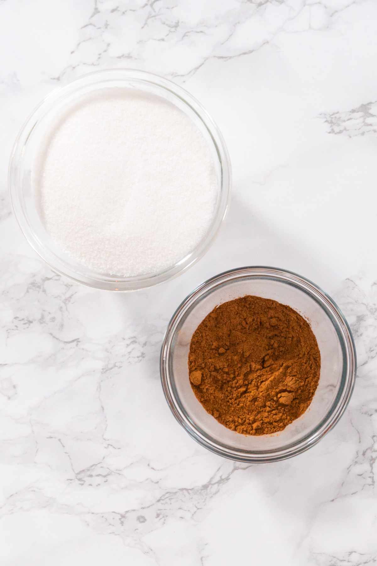 Two small glass bowls on a marble surface; one holds white granulated sugar and the other brown ground cinnamon, perfect for creating your own homemade cinnamon sugar recipe.