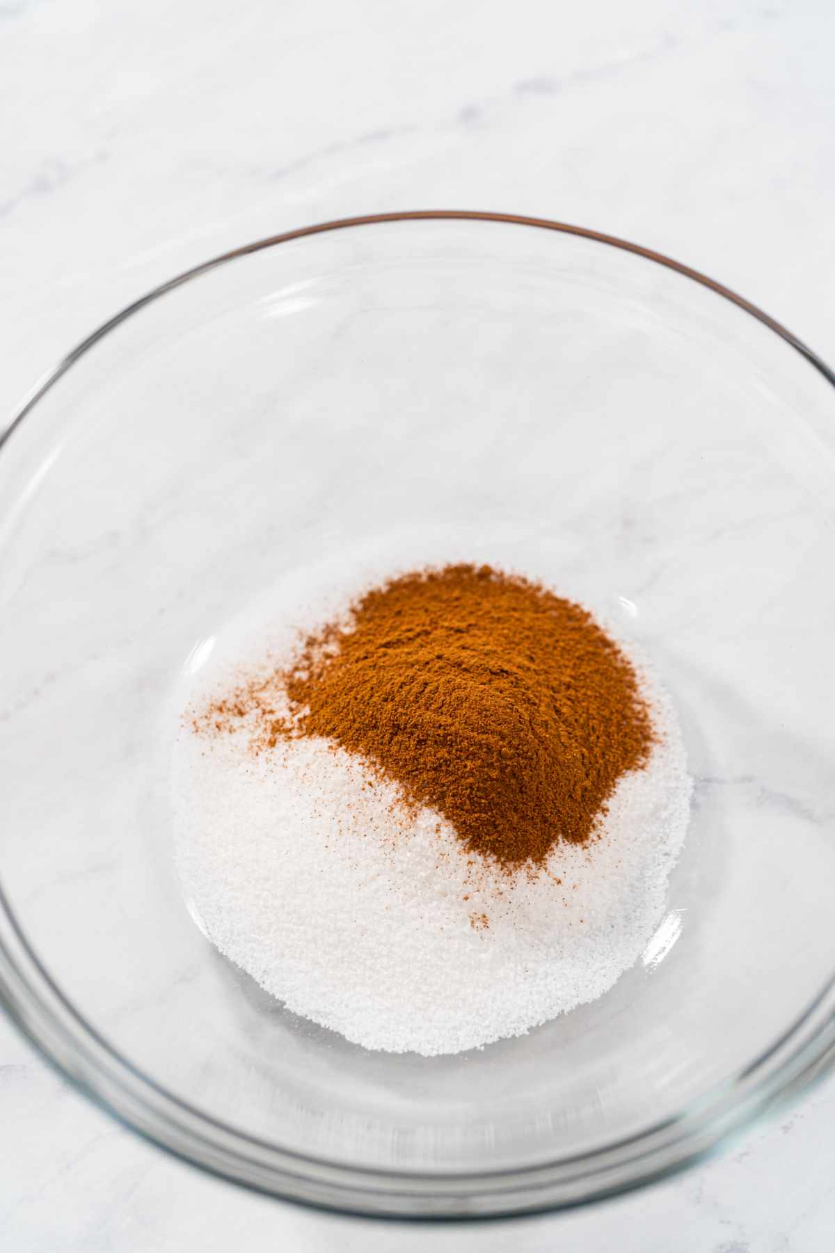 A glass bowl on a white surface contains a mound of white granulated sugar with a heap of brown cinnamon powder on top-perfect for a homemade cinnamon sugar recipe.