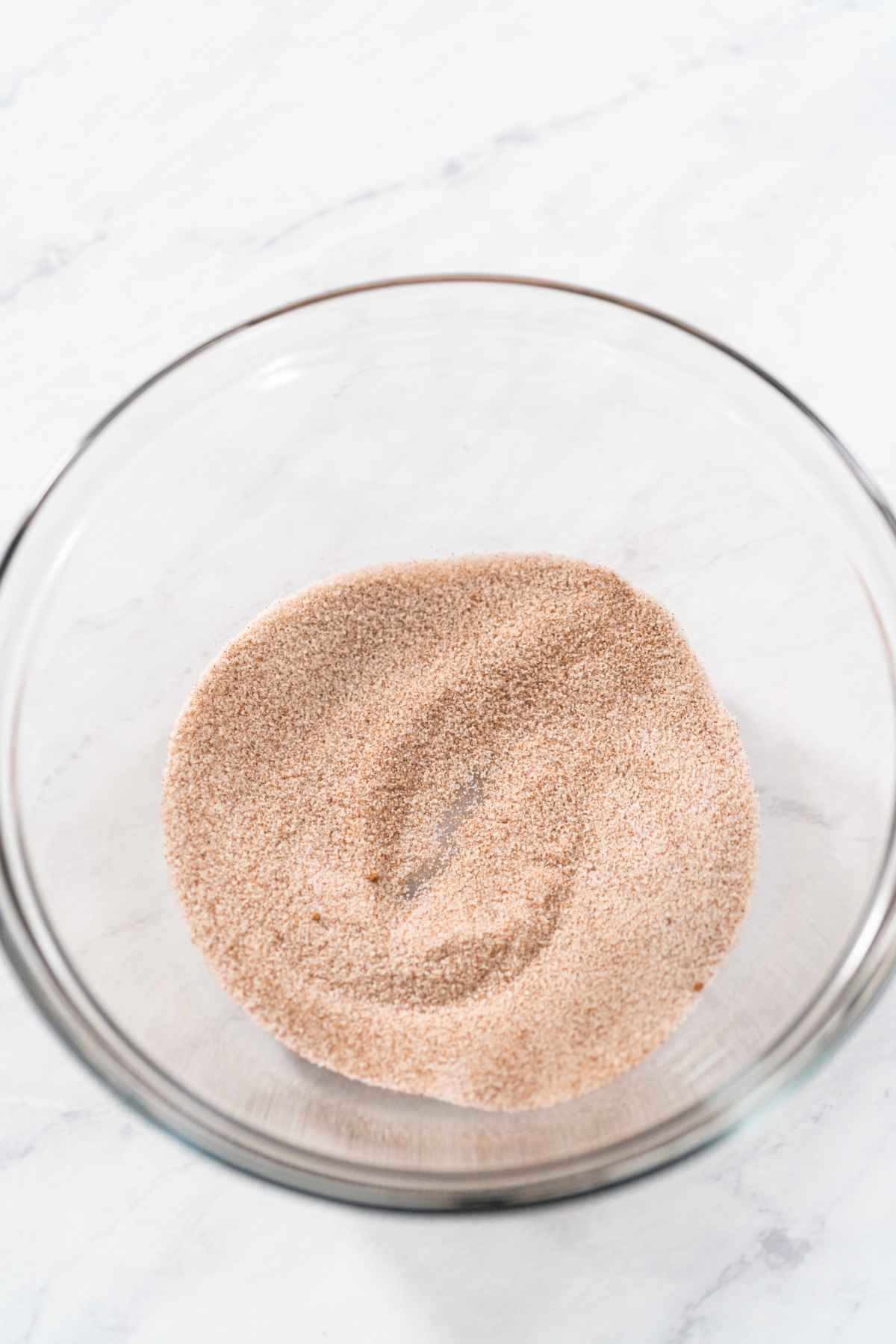 A glass mixing bowl containing a homemade blend of light brown granulated sugar and cinnamon, creating the perfect cinnamon sugar recipe, sits on a white marble surface.