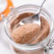 A close-up of a spoonful of homemade cinnamon sugar being held above a glass jar filled with more of the sweet mixture. The jar features a metal clasp and an orange seal.