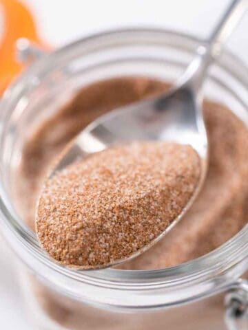 A close-up of a spoonful of homemade cinnamon sugar being held above a glass jar filled with more of the sweet mixture. The jar features a metal clasp and an orange seal.