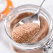 A spoonful of homemade cinnamon sugar held above a glass jar filled with the same mixture, with an orange lid visible in the background.