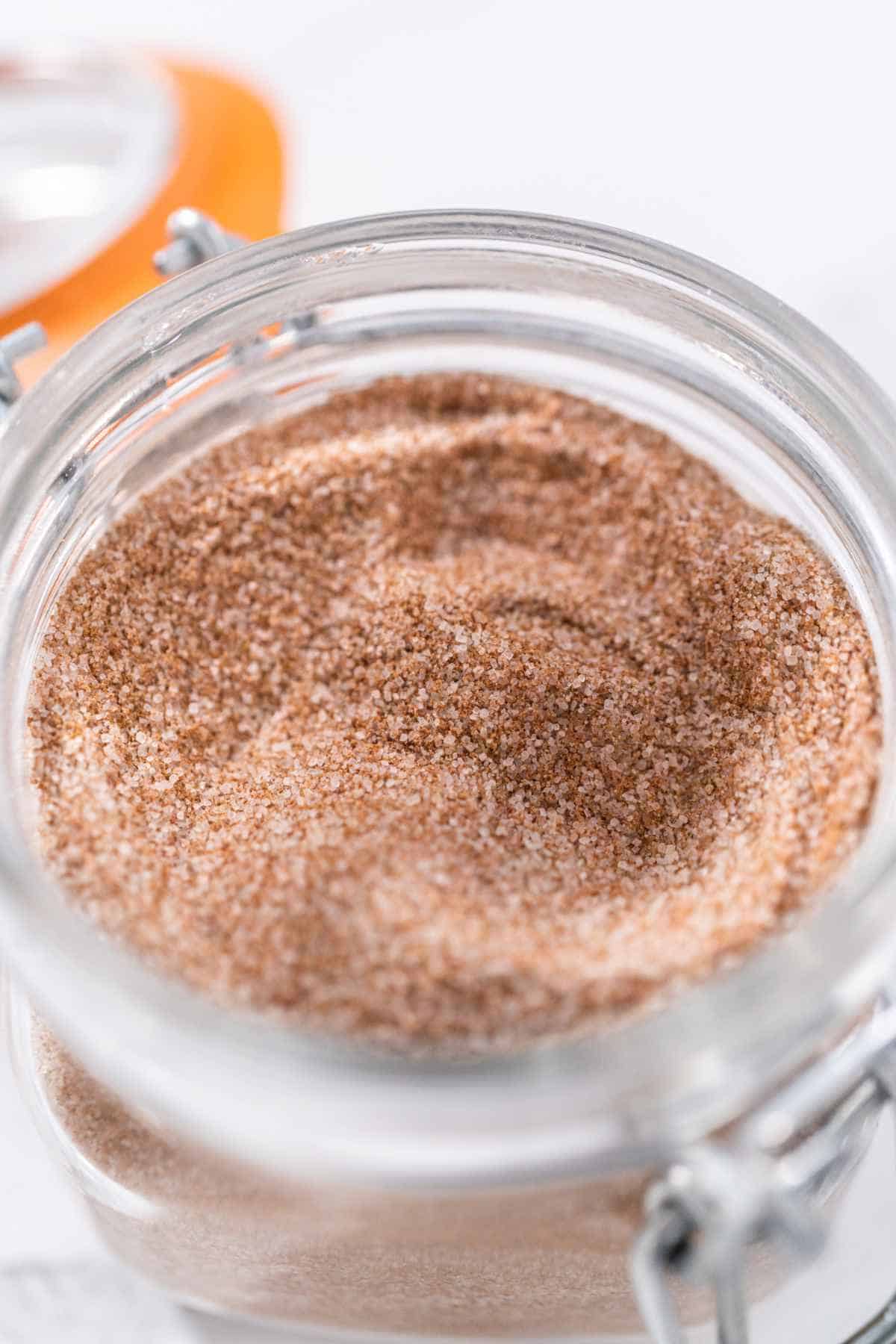 A close-up view of a glass jar filled with fine, light brown powder, likely a Cinnamon Sugar Recipe. The jar has a metal clasp and an orange rubber seal visible in the background.