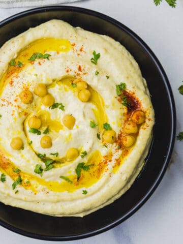 A black bowl filled with classic hummus dip, topped with whole chickpeas, a drizzle of olive oil, chopped parsley, and a sprinkle of paprika, on a light background.