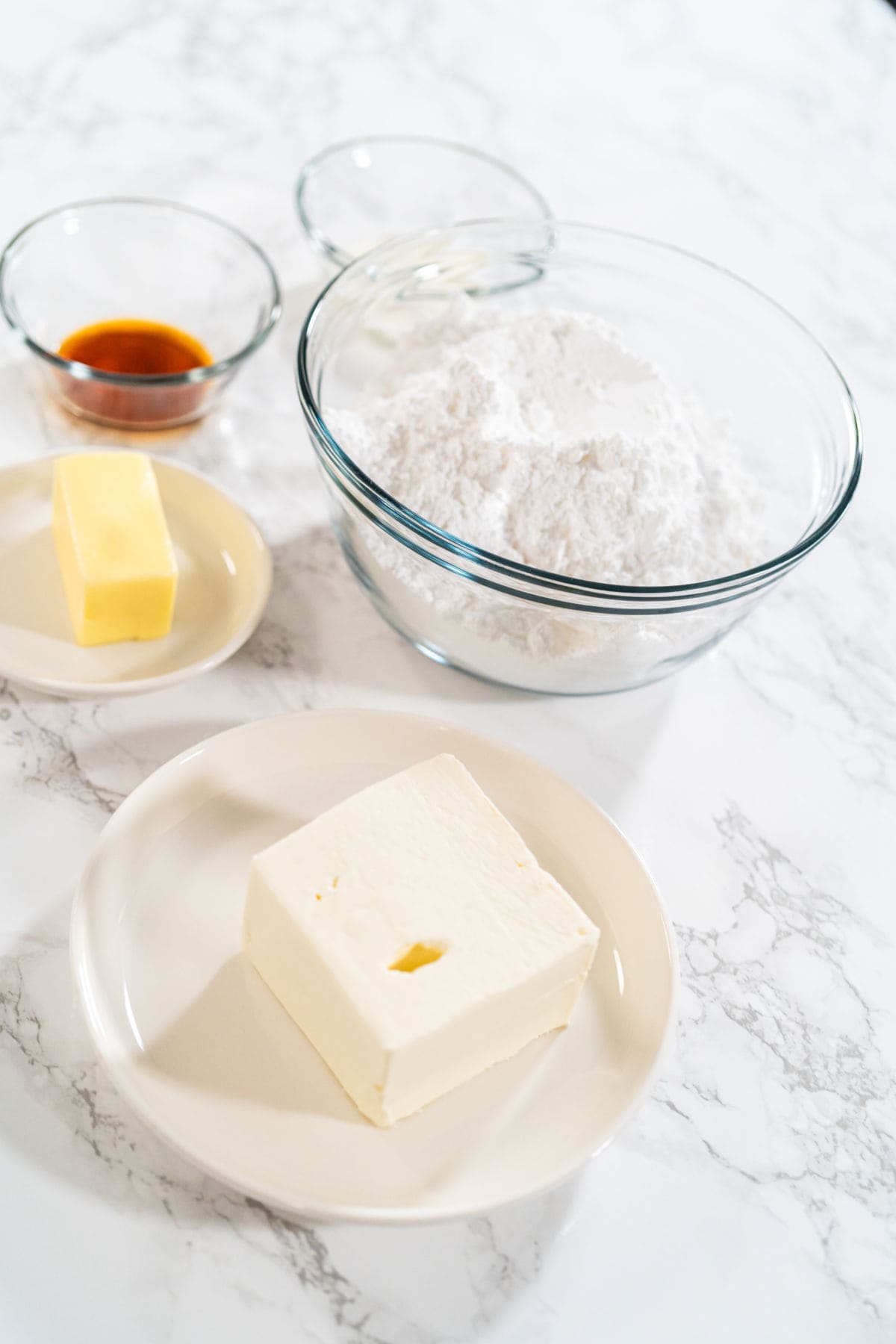A bowl of powdered sugar, a plate with cream cheese for cream cheese frosting, butter, and two small bowls with vanilla extract and milk are arranged on a white marble surface.