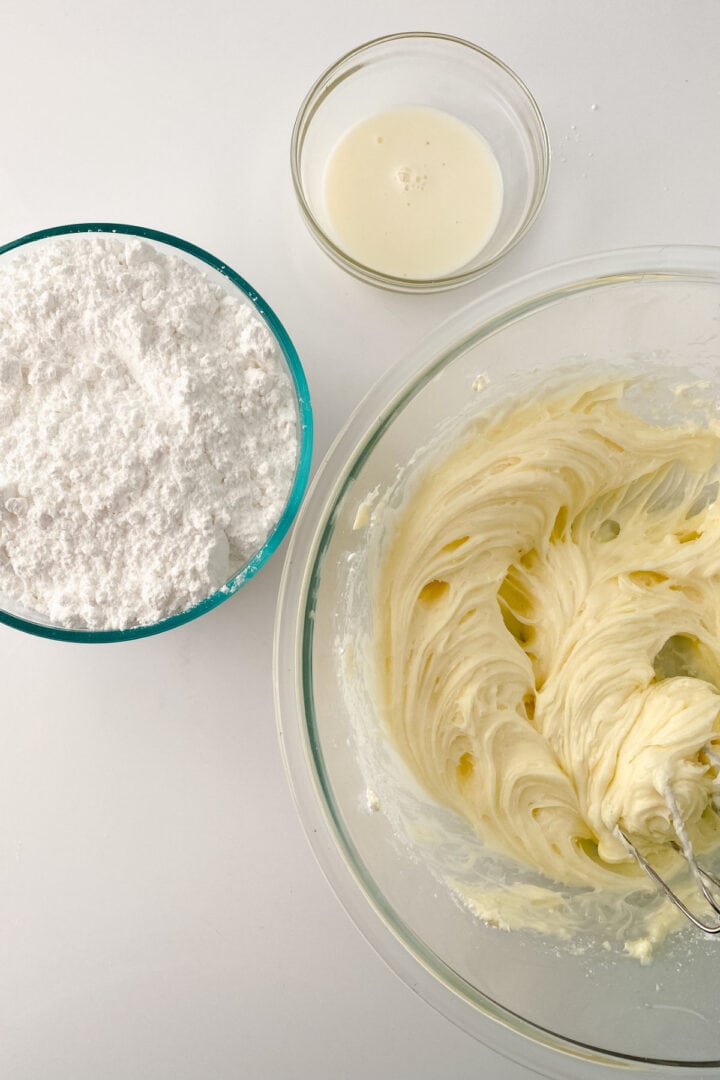 A glass bowl of whipped cream cheese frosting, a bowl of powdered sugar, and a small bowl of milk are arranged on a white surface. An electric mixer beater is partially visible in the frosting.