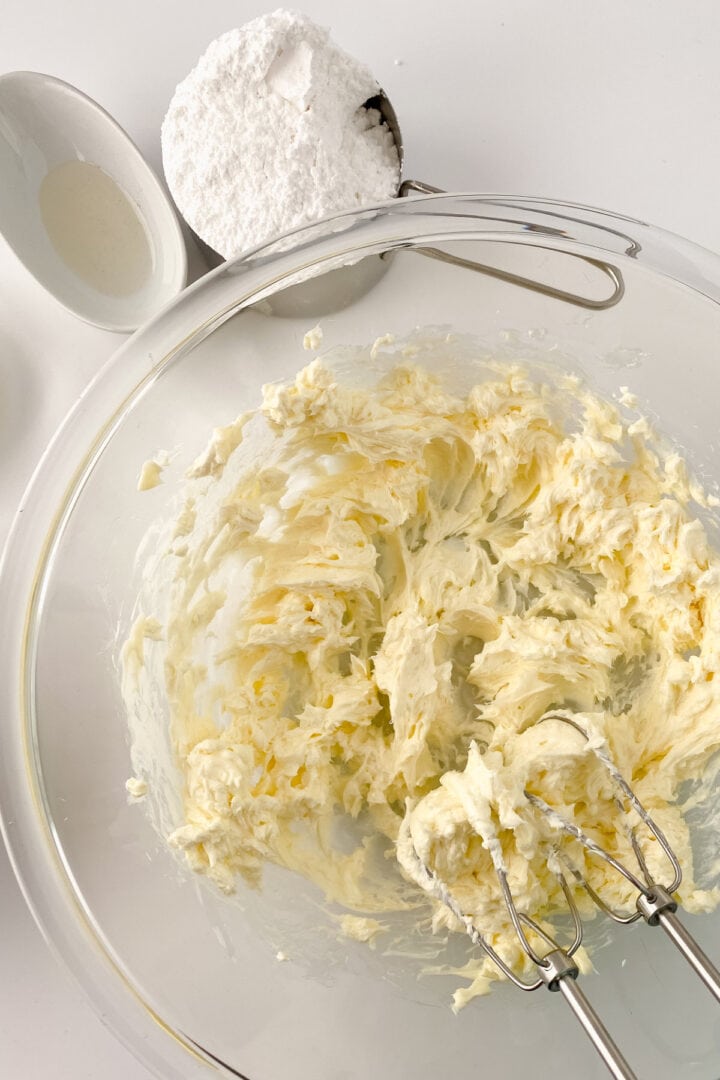 A glass bowl with creamed butter and sugar being mixed by electric beaters, perfect for making cream cheese frosting. Nearby are a metal measuring cup of powdered sugar and a small white bowl of liquid on a white surface.