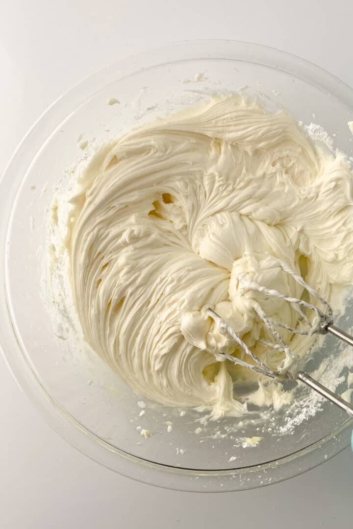 A glass bowl filled with smooth, creamy white cream cheese frosting is being mixed with a handheld electric mixer on a white surface.
